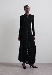 Kvinde iført en lang sort strikket kjole med knapper og lange ærmer samt sorte hæle, stående mod en simpel lys grå væg.