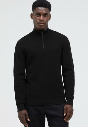 Schwarzer Strickpullover mit hohem Kragen und halb offenem Reißverschluss. Verfügt über eine gerippte Struktur und lange Ärmel, kombiniert mit dunkler Hose.