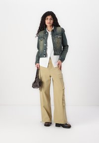 Giacca di denim con sfumatura blu-verde, camicia bianca, pantaloni cargo larghi color khaki, stivaletti alla caviglia marroni e una piccola borsa a tracolla marrone.