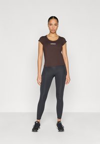 Dark brown fitted T-shirt with short sleeves and white "adidas" logo. Black high-waisted leggings with a smooth texture and tapered legs. Black sneakers.