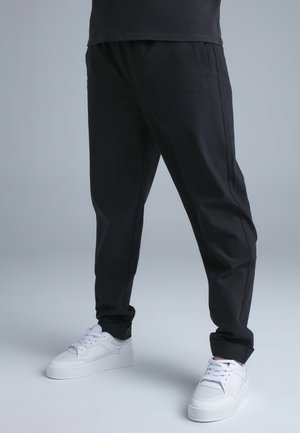 Personne portant un pantalon noir fuselé et des baskets blanches debout sur un sol gris clair devant un fond gris.