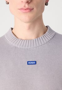 Gros plan d'une personne portant un pull côtelé gris clair avec un petit écusson bleu "HUGO" sur la poitrine et des boucles d'oreilles créoles argentées.