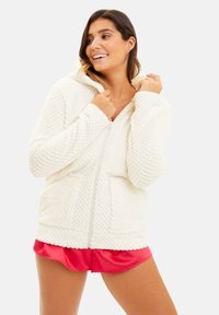 Veste à zip en polaire blanche avec un motif texturé, poches avant spacieuses et capuche. Portée avec des shorts en satin rose.