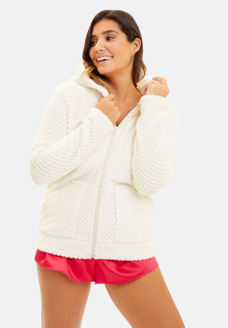 Veste à zip en polaire blanche avec un motif texturé, poches avant spacieuses et capuche. Portée avec des shorts en satin rose.
