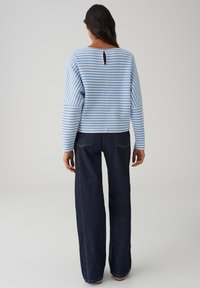 Maglione a righe blu e bianche con dettaglio a occhiello sul retro, abbinato a jeans a gamba larga di colore blu scuro. Il tessuto sembra morbido e strutturato.