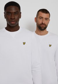 White long-sleeve sweatshirts with round necks; each features a small yellow bird logo on the left chest. The fabric appears soft and smooth.