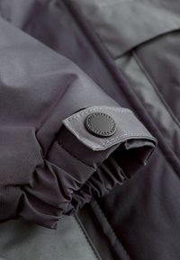 Poignet de veste grise avec tissu froncé et un bouton rond noir portant l'inscription "Sustainably Supplying". Texture lisse, fini mat, design minimaliste.