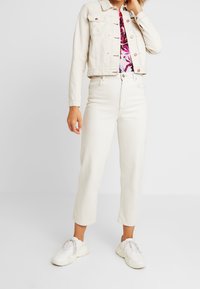 Chaqueta de denim blanca con botones de cobre, combinada con una blusa de estampado floral, jeans blancos de cintura alta y zapatillas blancas con suelas texturizadas.