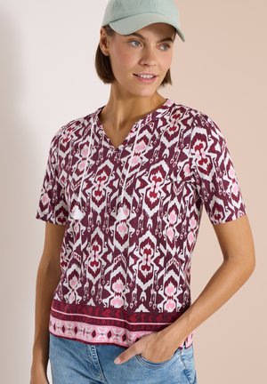 Kobieta ubrana w wzorzystą bluzkę z krótkim rękawem z frędzlami i jasnoniebieską czapkę z daszkiem, ręka w kieszeni, patrzy w prawo.