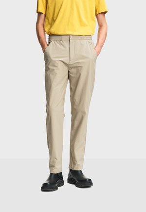 Homme portant un pantalon beige slim, une chemise à manches courtes jaune et des bottes noires épaisses, debout les mains dans les poches.