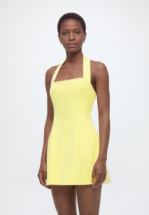 Kvinne med kort hår iført ermeløs gul halterhalskjole, stående mot ensfarget hvit bakgrunn.