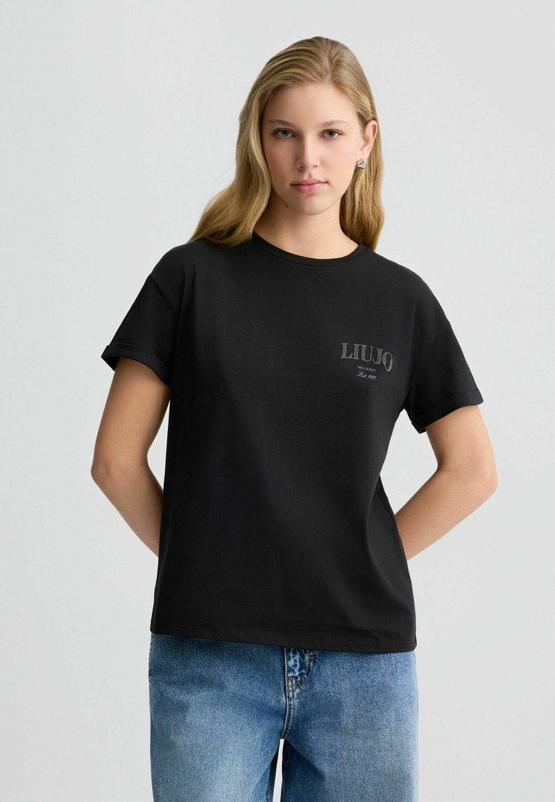 T-shirt noir à manches courtes en tissu lisse, avec un petit logo blanc sur la poitrine gauche. Associé à un jean en denim bleu clair.