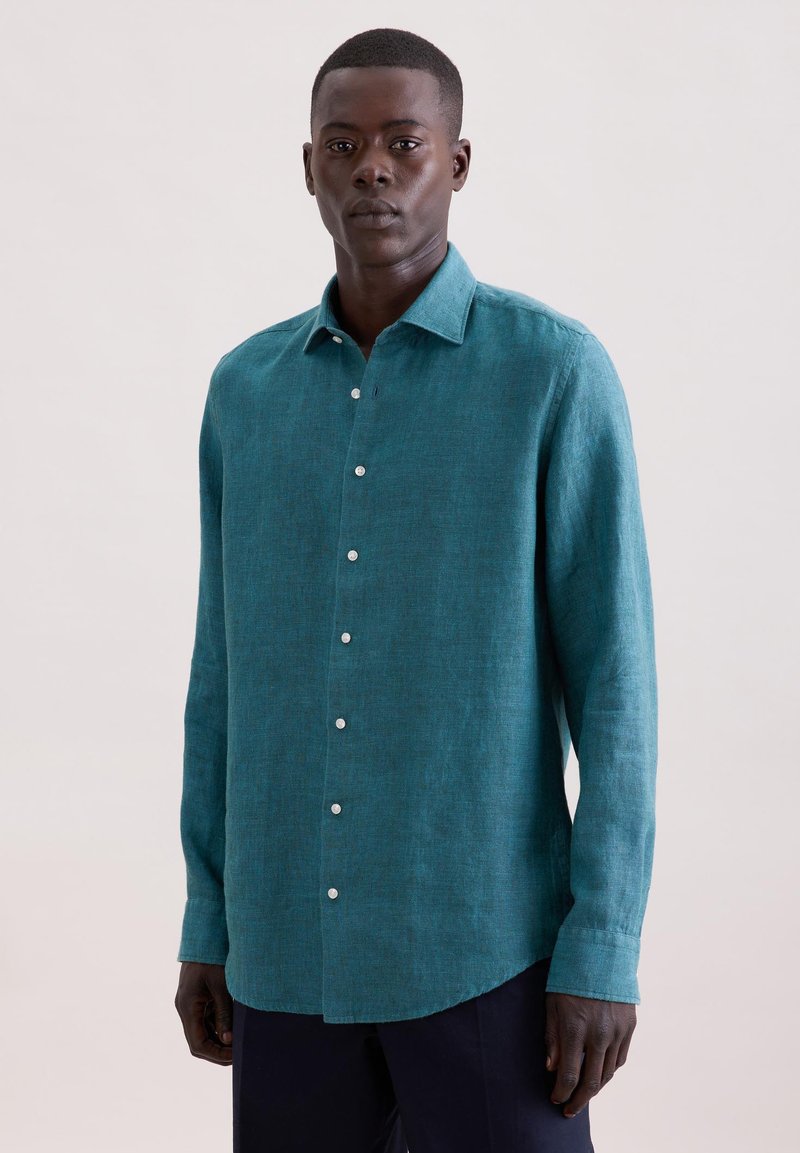 Homme à la peau foncée portant une chemise boutonnée à manches longues de couleur sarcelle et un pantalon sombre, debout devant un fond clair uni.