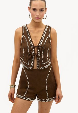 Femme portant un gilet sans manches en daim marron et un short assorti, tous deux ornés de clous argentés et d'un laçage devant, accessoirisée de boucles d'oreilles créoles et de bracelets.