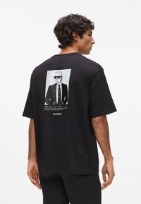 Črna bombažna majica s portretnim grafikonom na hrbtni strani, ki prikazuje osebo z sončnimi očali in obleko, z napisom pod sliko.