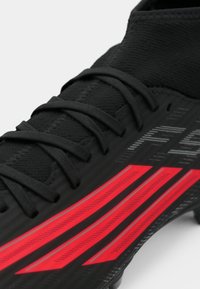 Μαύρο αθλητικό παπούτσι με υφή στο επάνω μέρος, διακοσμημένο με κόκκινες διαγώνιες ρίγες και παραδοσιακό κορδόνι. Η κοντινή γωνία προσφέρει έμφαση στις λεπτομέρειες του σχεδιασμού.