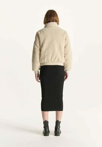 Giacca fuzzy beige con collo alto, indossata sopra una gonna midi aderente nera in maglia e stivaletti neri, vista da dietro.