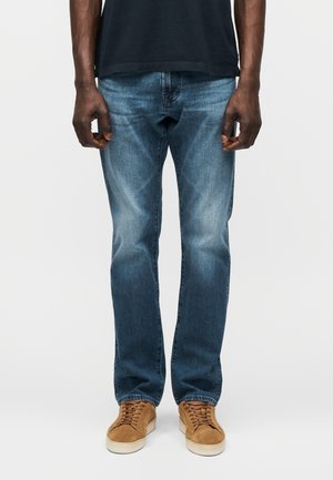 Mand iført falmede blå jeans, brune ruskindssneakers og en mørk skjorte, stående mod en ensfarvet lys baggrund.