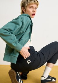 Teal zip-up hoodie over white shirt, black pants with logo patch, black sneakers with white stripes, and white cotton socks on a yellow floor.