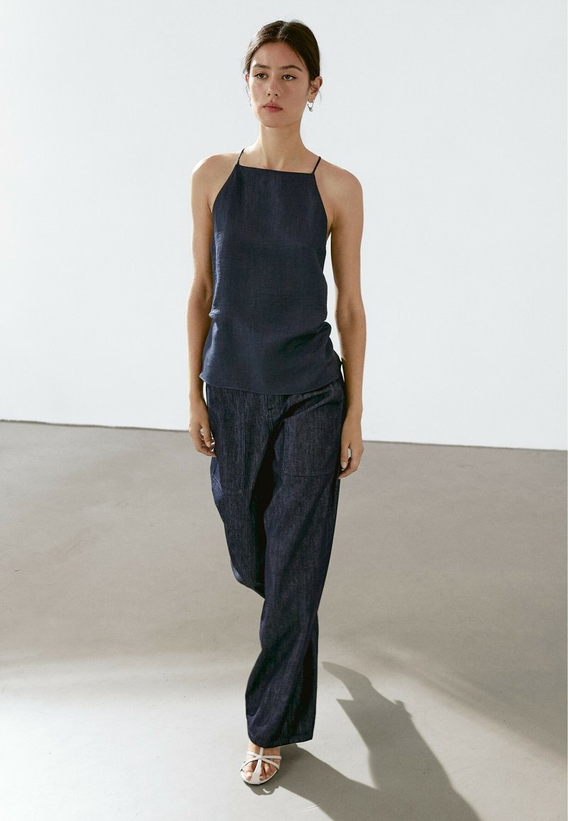 Top senza maniche di colore blu navy con spalline sottili, abbinato a pantaloni larghi coordinati. Texture liscia con un leggero luccichio. Scarpe bianche con punta aperta.