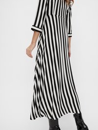 Maxi obleka z vertikalnimi črnimi in belimi črtami, s tremi četrtinami rokavi, prilegajočim zgornjim delom in prosto padajočo krilom. Poudarjena z bleščečimi čevlji.
