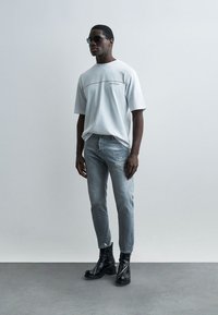 Άνδρας που φοράει λευκό υπερμεγέθες T-shirt, ανοιχτό γκρι τζιν με φθορές, μαύρες μπότες με τακούνι και μαύρα γυαλιά ηλίου, στέκεται σε απλό τοίχο.