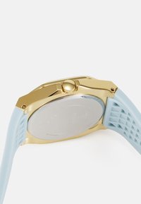Montre avec boîtier doré et bracelet en silicone bleu présentant un motif texturé. Le dos de la montre rond montre des détails gravés.