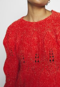 Un pull en tricot rouge présente un motif ouvert avec une finition texturée, un col rond et des motifs distincts en forme de feuille sur l'ensemble.