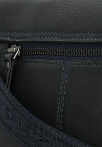 Borsa in pelle nera con zip metallizzata, cuciture blu scuro e tracolla intrecciata con testo impresso. Primo piano dell'area della zip.
