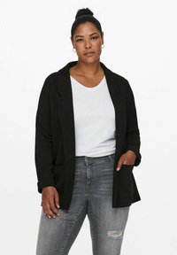 Černý blejzr s jemnou texturou, otevřený design, dvě přední kapsy, nošený přes bílou v-neck košili, spárován se šedými potrhanými džíny.