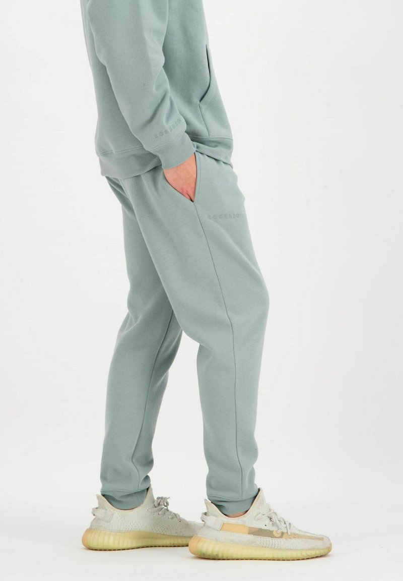 Hellgrüne Sweatpants aus weichem Material, mit Seitentaschen und gerippten Bündchen, kombiniert mit hellbeigen Sneakers.