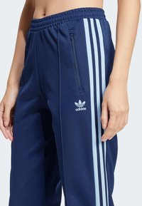Detailný záber osoby nosiacej tmavomodré teplákové nohavice Adidas s bledomodrými pruhmi na boku, vreckom na zips a logom pri páse.