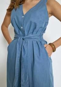Chambray haljina s V-izrezom, prednjim gumbima, pojasom u struku i bočnim džepovima. Svijetloplava tkanina s glatkom teksturom. Smeđi narukvica na zapešću.