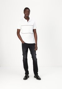 Hvid polo skjorte med en vandret burgunderstripe, parret med sorte jeans og sorte sko. Modellen står foran en hvid baggrund.