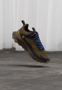 Brauner und schwarzer Wanderschuh mit blauen Schnürsenkeln, der über einem grauen Betonboden und einem Hintergrund aus transparentem weißen Vorhang schwebt.