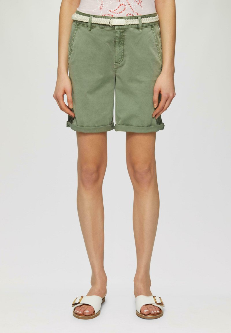 Shorts vert clair arrivant aux genoux avec des revers, dotés de poches avant et d'une ceinture en tissage crème. Portés avec des sandales blanches.