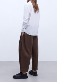 Camicia bianca a maniche lunghe con sottili righe verticali, abbinata a pantaloni larghi marroni e sneaker nere. Tasche visibili sui pantaloni.