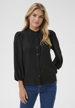 Blouse noire en tissu texturé avec un col montant, des détails à volants et une fermeture à boutons. Dispose de manches bouffantes et d'une coupe décontractée.