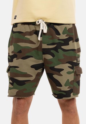 Short cargo camouflage pour homme avec poches latérales et taille à cordon blanc, porté avec un haut beige.