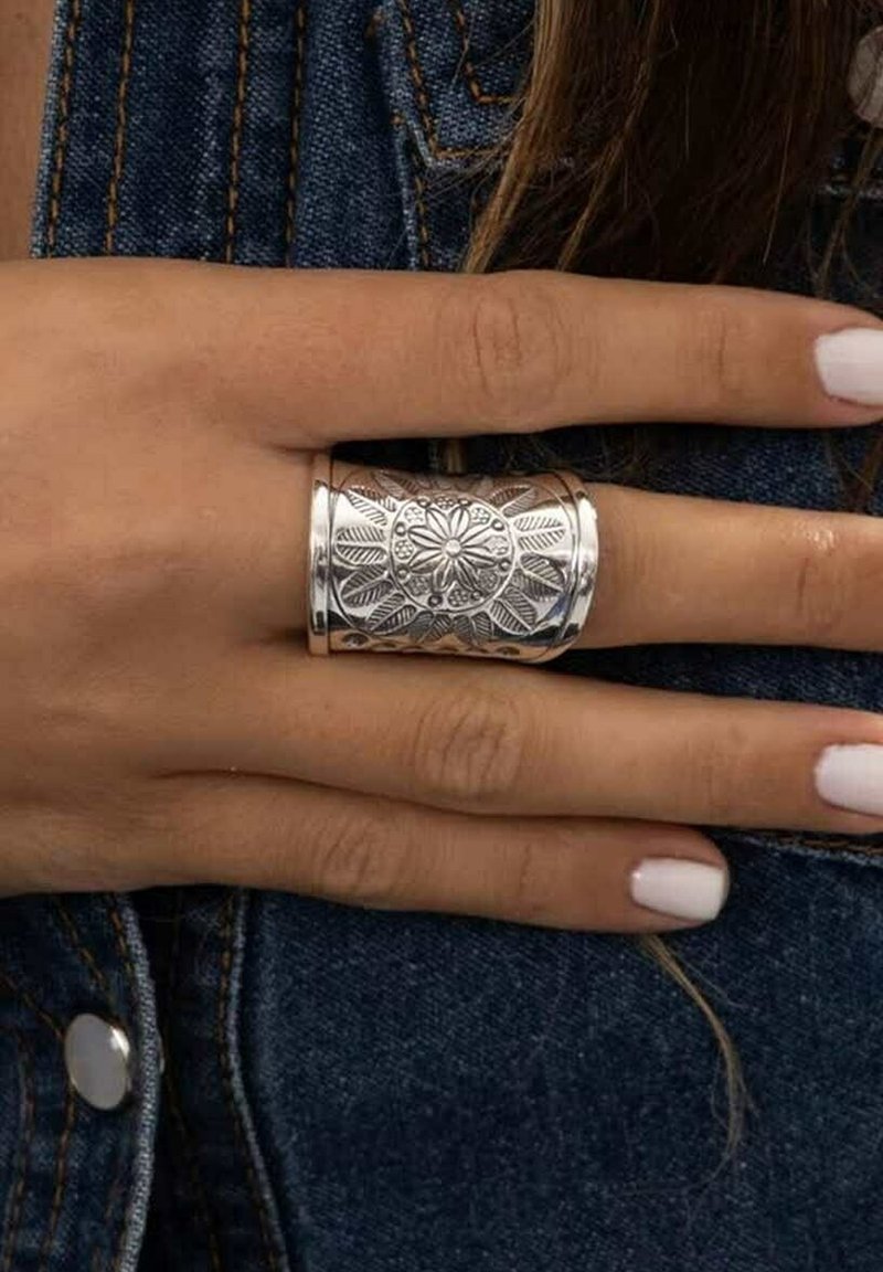 Main avec des ongles peints en blanc portant une large bague en argent gravée de motifs floraux et de feuilles, reposant sur un tissu en denim.