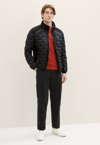 Giacca imbottita nera con colletto alto e materiale a contrasto sulle spalle, indossata sopra una camicia rossa, abbinata a pantaloni neri e sneakers bianche.