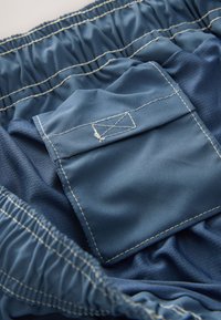 Plava tkanina glatke teksture s nabranim pojasom; sadrži zakrpni džep s vidljivim detaljima šavova.