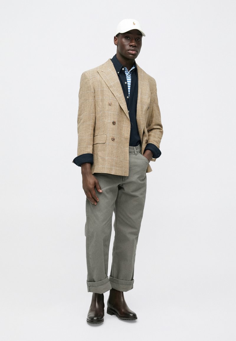 Beige plaid double-breasted blazer, navy polo with stripes, gray rolled-up chinos, and brown boots. Accompanied by a white cap.