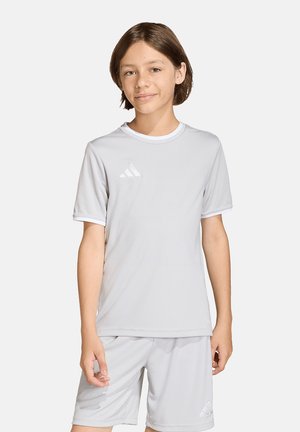 Junge Person mit kurzem braunem Haar, die ein hellgraues Adidas-Sport-T-Shirt und passende Shorts trägt, vor einem schlichten Hintergrund stehend.