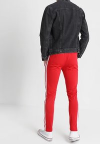 Černá džínová bunda se rovný střih, spárovaná s červenými slim-fit kalhotami se bílými bočními pruhy. Bílí tenisky dokončují outfitu.