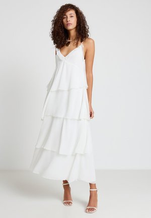 Femme aux cheveux bouclés portant une robe longue blanche sans manches à étages et des talons blancs à bride à la cheville, debout devant un fond uni.