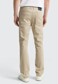 Beige Baumwollhosen mit geradem Schnitt, ausgestattet mit zwei Gesäßtaschen und einem dunklen Stoffetikett über der rechten Tasche. Getragen mit weißen Sneakers.