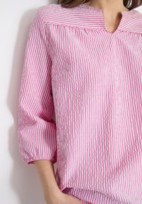 Mujer con blusa de manga larga de rayas verticales en rosa y blanco, con escote en forma de V y puños elásticos en las mangas.