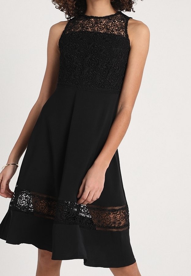 Robe noire sans manches longueur genou avec des détails en dentelle florale sur la poitrine et au-dessus de l'ourlet, portée par une personne aux cheveux bouclés.