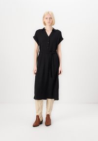 Rochie neagră cu mâneci scurte, cu talie strâns�ă de o curea, până la genunchi, fabricată dintr-un material texturat. Modelul poartă botine maro până la gleznă.